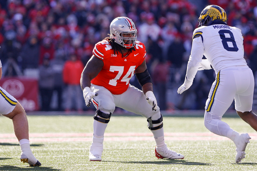 Donovan Jackson (red jersey, white #74, white pants, high white socks, prepares to block a Michigan player.