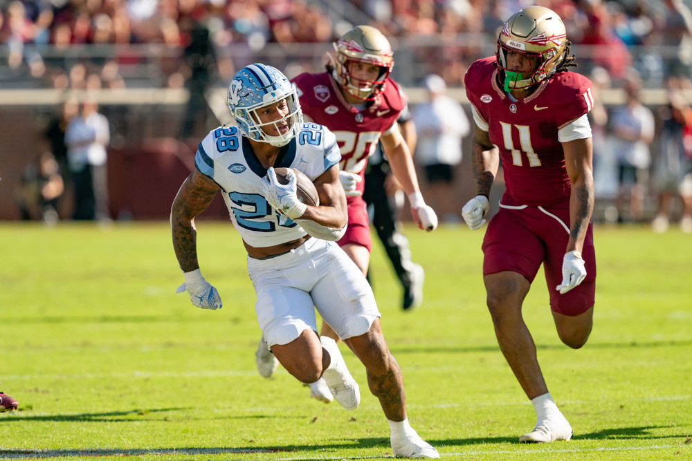 Omarion Hampton of North Carolina sprints away from a Florida State player. He's wearing a white uniform with light blue trim and numbers.