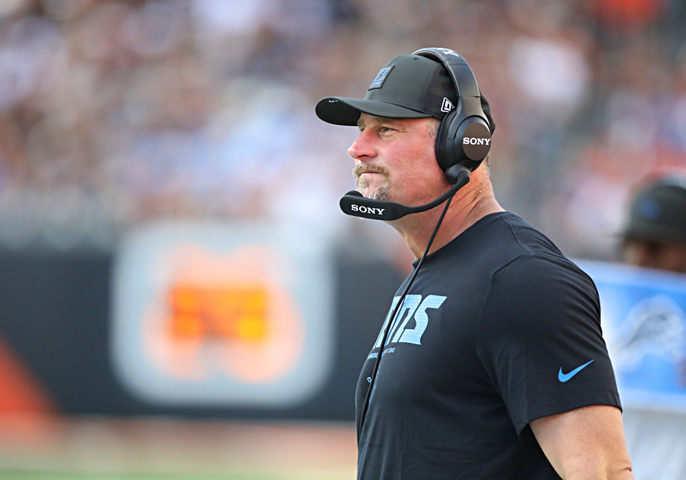 Headset-wearing Lions coach Dan Campbell watches from the sidelines. He's wearing a Lions t-shirt.
