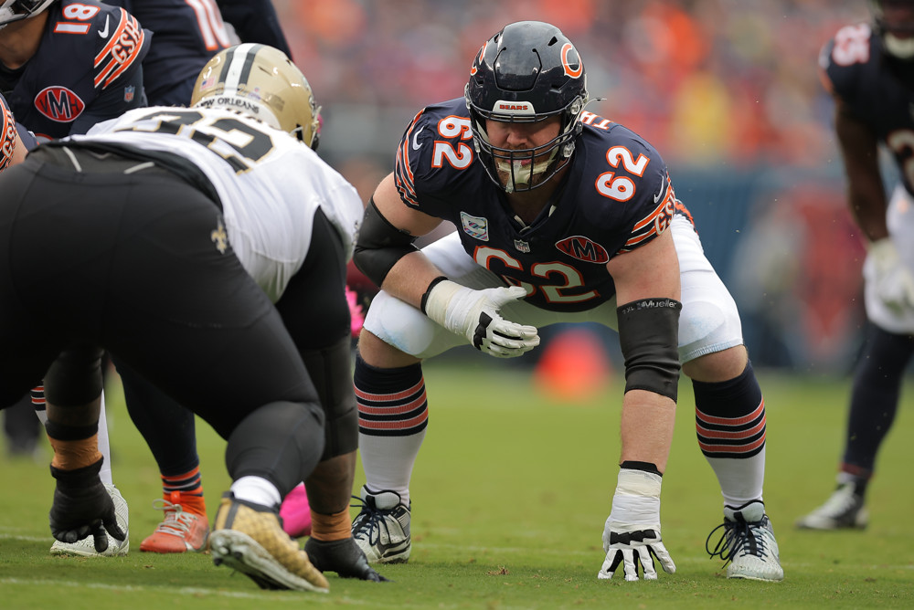 Joe Thuney - wearing a black jersey with #62, bends over, with 4 fingers on his left hand touching the turf and his right hand close to his chest as he prepares for the football to be snapped.