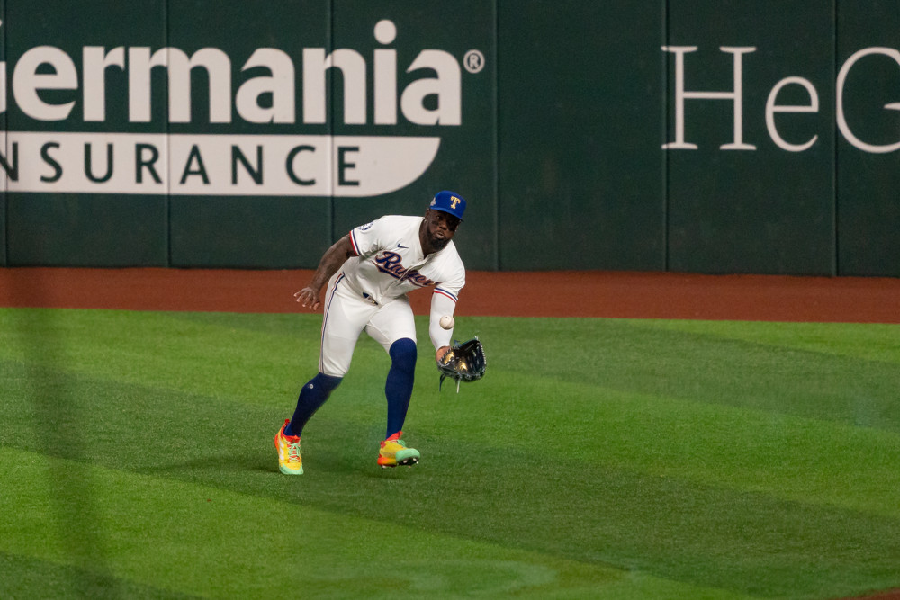 Adolis Garcia, in white Rangers jersey with blue cap, high blue socks and red/yellow/green colored sneakers, catches a ball knee high with his glove.