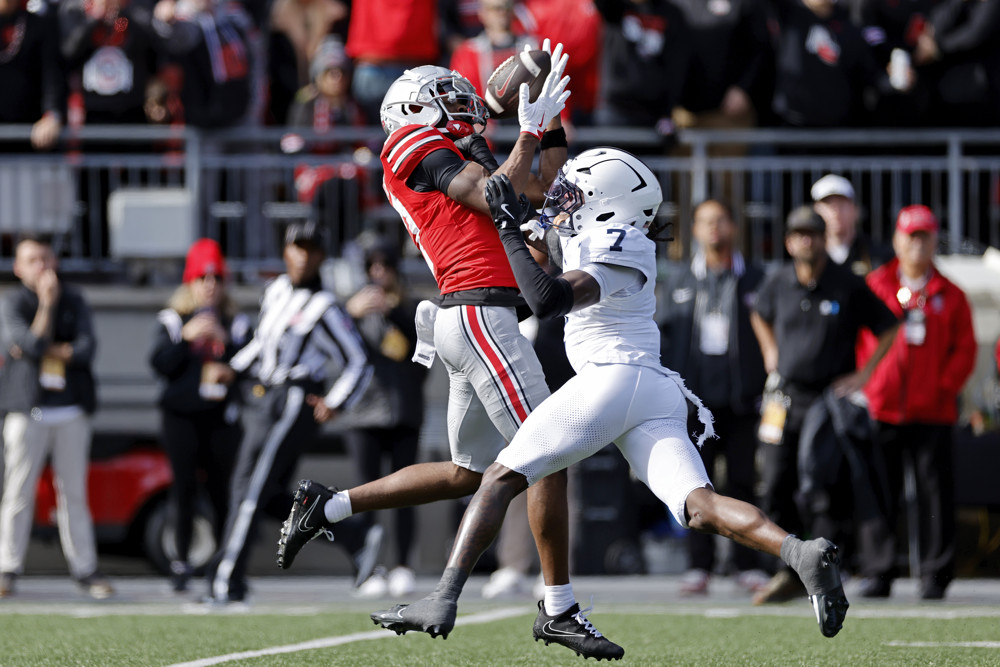 Carnell Tate, wearing a red Ohio State jersey and grey pants, reaches up over a defensive player who is facing him (wearing an all-white uniform) to make a catch.
