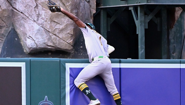 Denzel Clarke, wearing a grey Athletics jersey and pants, reaches high over the center field fence with the glove on his left hand to make a catch. He's propping himself up higher using his right hand.