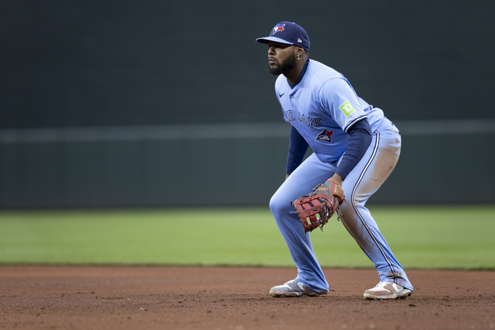 Vladimir Guerrero Jr. wearing light blue pants and a light blue Blue Jays jersey and dark blue Blue Jays cap, has his glove on his left hand and facing outward as he waits for a pitch to be thrown.