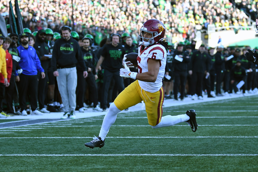 A player in a USC helmet, a white jersey, yellow pants, and high white socks, runs toward the end zone with the football in his right hand.