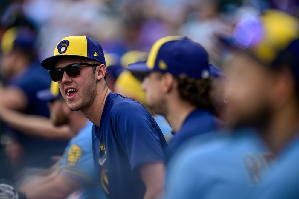 A group of players on the Milwaukee Brewers sit in the dugout and look to their right, among them pitcher Jacob Misiorowski who is wearing a yellow cap with the Brewers logo and sunglasses.
