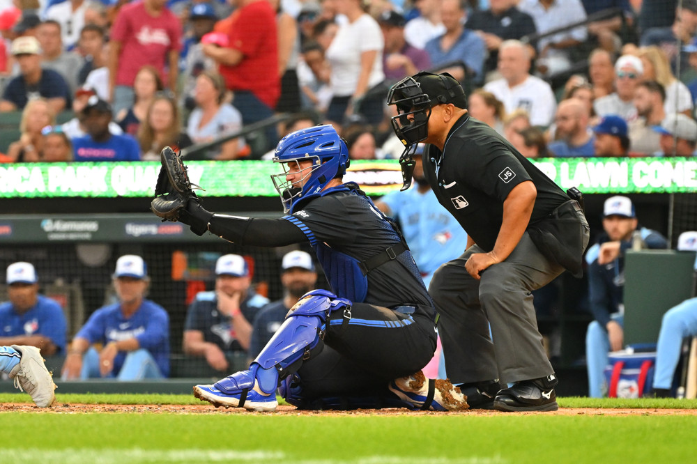Dillon Dingler crouches and stretches out his right leg, and holds his glove out as he prepares to catch a pitch with the umpire crouching behind him.