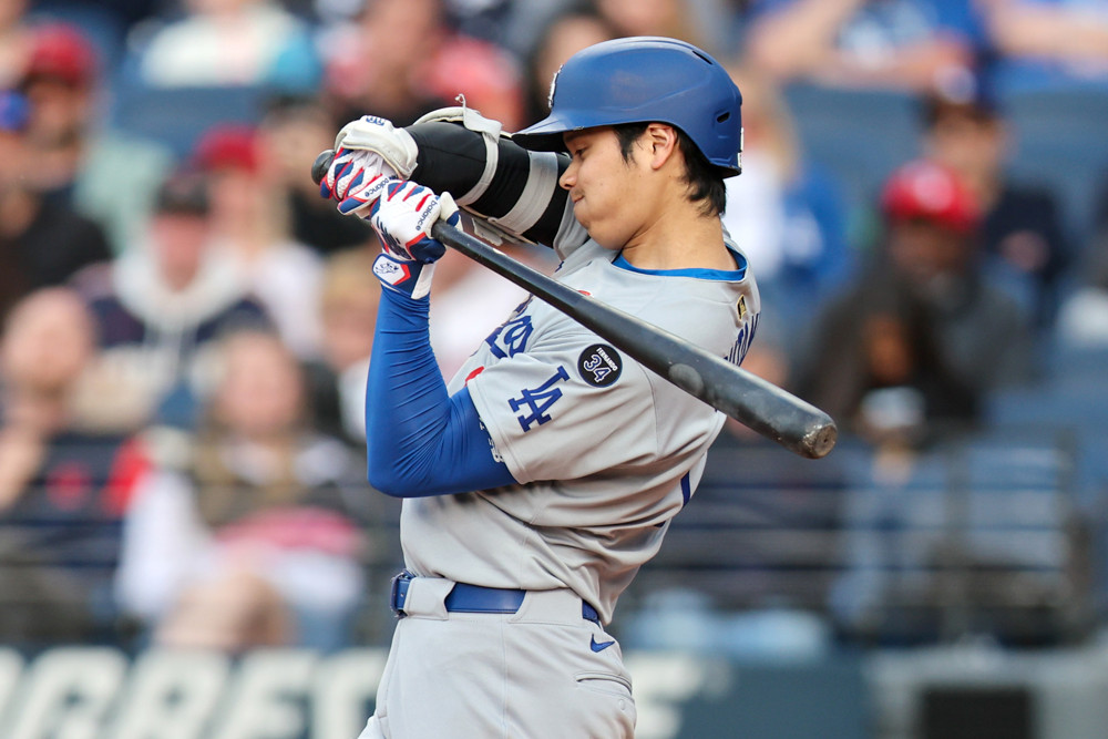 Shohei Ohtani winces as he starts to swing the bat, then stops. He is wearing a blue helmet and a grey Los Angeles Dodgers jersey.