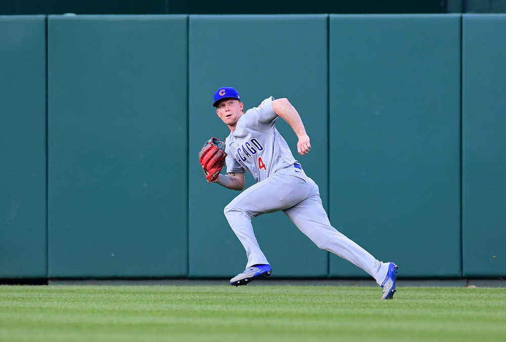 Pete Crow Armstrong, wearing a grey Cubs jersey and blue Cubs cap, retreats to try to catch a fly ball near the center field fence.